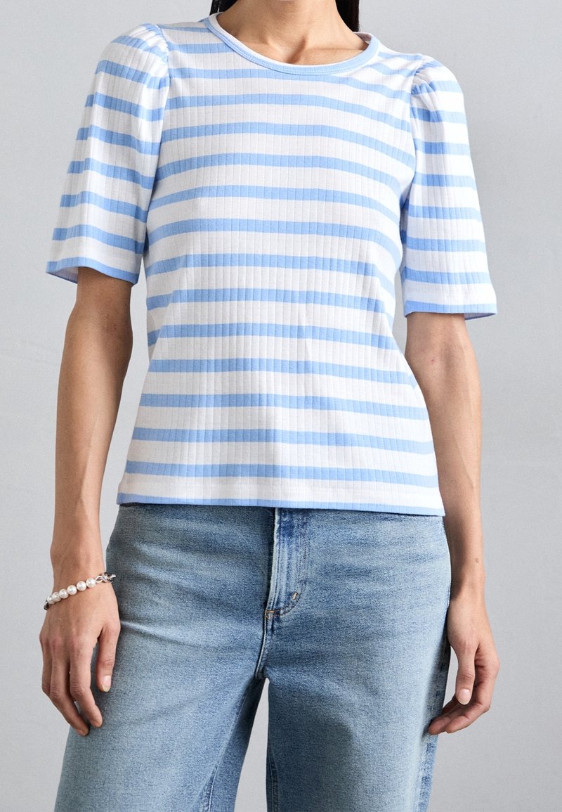Femme portant un haut à manches courtes rayé horizontalement bleu clair et blanc avec des épaules bouffantes et un jean en denim bleu clair.