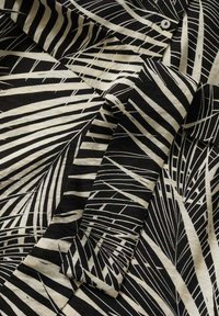 Tissu noir et beige avec un motif abstrait de feuilles, montrant des manches pliées et un bouton blanc près du col.