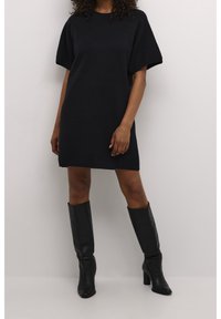 Femme portant une robe noire à manches courtes associée à des bottes en cuir noires à talons montant jusqu'aux genoux, debout devant un fond blanc uni.