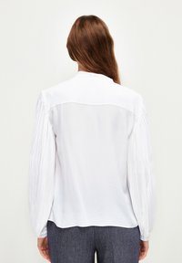 Weiße Bluse mit plissierten langen Ärmeln, glatter Textur, klassischem Kragen und lockerer Passform. Die Rückansicht zeigt die Schlichtheit des Designs.