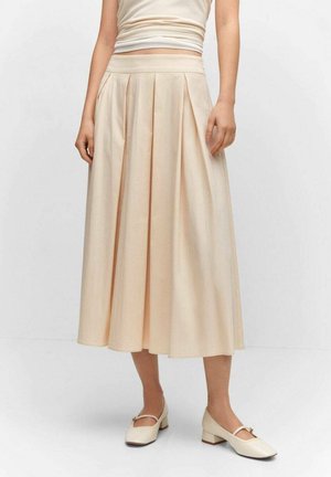 Femme portant une jupe midi plissée beige taille haute avec des chaussures Mary Jane à talon carré nude, debout devant un fond clair uni.