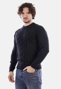 Uomo con capelli ricci che indossa un maglione nero lavorato a trecce e jeans blu, in piedi con una mano in tasca contro uno sfondo bianco.