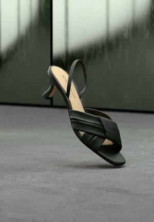 Black leather slingback sandal with woven straps and low kitten heel, displayed against a blurred dark gray background.