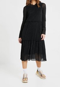Svart långärmad klänning med en texturerad, glittrig tyg, med en volangkjol och en scallopad halsringning. Matchad med färgglada sneakers.