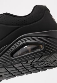 Black athletic shoe featuring a smooth upper, perforated detailing, and a rubber sole with a visible air cushioning unit and textured tread design.