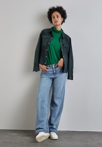 Tmavě zelená semišová košile, zelený rolák, světle modré široké džíny, bílé tenisky, postávající proti obyčejné zdi.