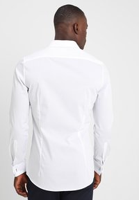 Hvit langermet skjorte laget av glatt stoff, med klassisk krage, knappemansjetter og buet nederkant.