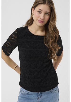 Mladá žena s dlhými vlnitými vlasmi, ktorá má na sebe zlaté kruhové prívesky, čierny čipkovaný krátky rukáv, modré džínsy a zlatý náramok, pózujúca s rukami za chrbtom.