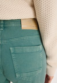 Jeans de mezclilla verde con un acabado de tela texturizada. Presenta un bolsillo trasero y una etiqueta de cuero marrón con letras en relieve.