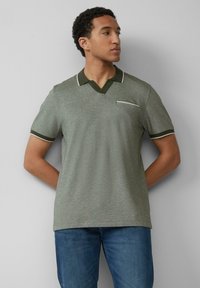 Polo grigio-verde con scollatura a V, colletto verde scuro a contrasto e rifiniture bianche. Presenta una piccola tasca sul petto. Abbinato a jeans blu.