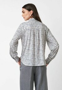 Frau mit langen dunklen Haaren, die Perlenohrringe und eine weiße Bluse mit einem schwarzen Karomuster trägt, kombiniert mit lockeren grauen Hosen, die von hinten zu sehen ist.