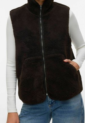 Personne portant une veste noire à fermeture éclair en matière pelucheuse par-dessus une chemise blanche à manches longues et un jean bleu, une main dans la poche de la veste.