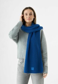 Femme jeune avec des cheveux bruns lisses portant un pull gris et un jean gris clair, ajustant une longue écharpe en tricot bleu autour de son cou.