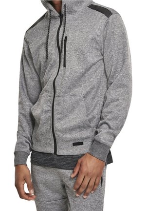Mann trägt grauen Kapuzenpullover mit Reißverschluss und schwarzen Akzenten, Fronttaschen und passende graue Jogginghose, steht mit einer entspannten Hand an der Seite.