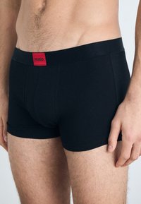 Schwarze Boxerbriefs mit glatter Textur, auf der der rote HUGO Logo am Bund prangt. Das Design ist enganliegend und reicht bis zur Mitte des Oberschenkels.