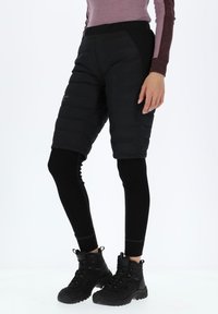 Zwarte geïsoleerde shorts over zwarte leggings, met een strakke uitstraling, gewatteerde textuur en elastische tailleband. Zwarte wandelschoenen zichtbaar.