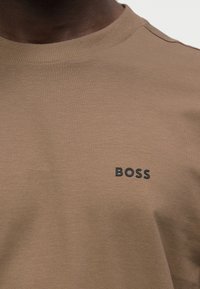 T-shirt en coton brun avec un col ras du cou côtelé. Présente un logo "BOSS" noir sur la poitrine. Le tissu a une apparence texturée et tissée.