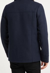 Tmavě modrý vlněný kabát se strukturovaným střihem, s vysokým límcem, švovými detaily a dlouhými rukávy, zobrazený zezadu.