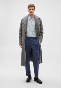 Γκρι μάλλινο παλτό, λευκό πουκάμισο με μπλε ρίγες, μπλε παντελόνια και μαύρα λοφάρια· εφαρμοστή γραμμή και απαλές υφές με λεπτομερείς πινελιές.