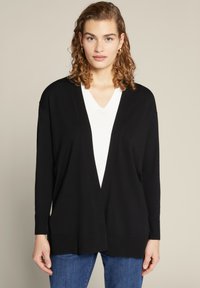 Cardigan nero con apertura davanti, maniche lunghe e vestibilità morbida. Indossato sopra una maglietta bianca a scollo a V. Tessuto liscio.