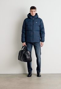 Marineblå tynd dunjakke med hætte, lynlåslukning og quiltet design; parret med mørk denim og en sort læder sportstaske. Glat tekstur.