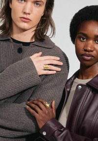 Cárdigan de punto acanalado marrón con un botón negro, llevado con un anillo dorado. La chaqueta de cuero oscura acentúa el detalle en una mano.