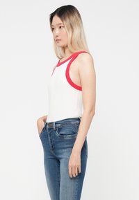 Camiseta sin mangas blanca con detalles rojos y amplios agujeros para los brazos, combinada con jeans de mezclilla azul. La camiseta tiene un diseño ajustado y una textura suave.