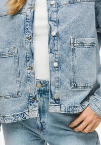 Hellblaues Jeansjacke mit silbernen Druckknöpfen, ausgestattet mit zwei Vorderaschen und einem leicht abgetragenen Aussehen. Trägt man über einem weißen Oberteil.