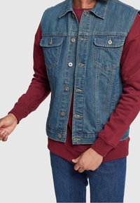 Denimväst med två bröstfickor, avslappnad passform, blå nyans, med metallknappar, sliten textur, lager på lager över en bordeauxfärgad sweatshirt.