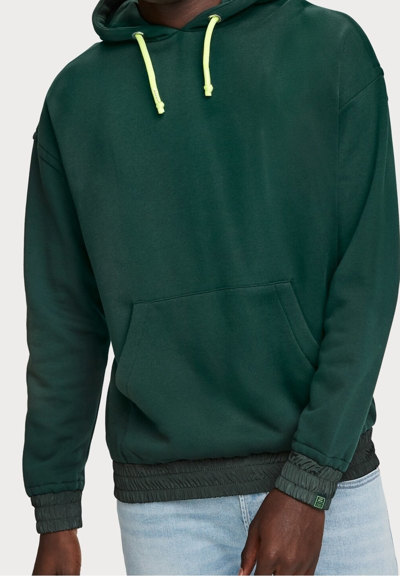Sudadera verde oscura con un bolsillo frontal, cordones ajustables amarillos y puños elásticos. Presenta un tejido texturizado con un color sólido.