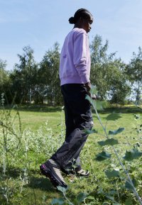 Felpa viola chiaro con logo sulla schiena, pantaloni cargo neri e scarpe da esterno multicolori. Circondato da erba e piccole piante.