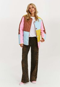 Žena v farebne kombinovanej prešívanej bunde, bielom tričku, širokých nohaviciach s leopardím vzorom a ružových teniskách stojí pred jednoduchým pozadím.