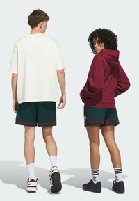 Two people from the back wearing sportswear: a white oversized t-shirt, maroon hoodie, green shorts with red trim, and various sneakers.