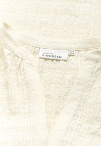 Blusa cremoso texturizzata con scollatura a V, dotata di un'etichetta che recita "KAREN BY SIMONSEN", realizzata in un tessuto leggero e intrecciato.