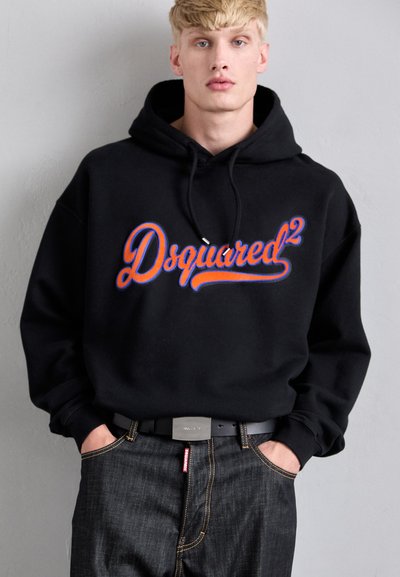 Mladý muž v černé mikině s tučným oranžovým a modrým logem "Dsquared2", tmavých džínách a černým páskem, stojící u světle šedé zdi.