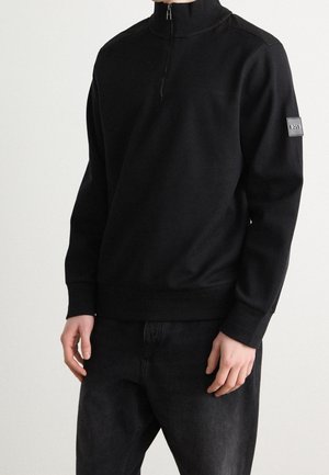 Pulóver negro con cuello con cremallera y textura acanalada, mangas largas y cintura ajustada. Presenta un parche de logo en la manga izquierda. Combinado con jeans oscuros.