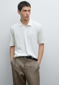 Maglietta polo bianca realizzata in materiale morbido con colletto classico e patta con tre bottoni, abbinata a pantaloni chiari in maglia marrone.
