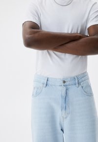 Jeans de mezclilla azul claro con un corte relajado, que cuentan con bolsillos frontales, un cierre de botón y una textura deslavada, usados con una camiseta blanca.