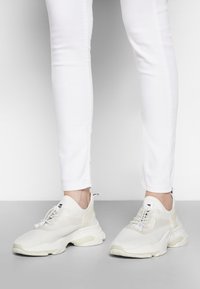 Scarpe sportive bianche con tomaia in rete, linguetta imbottita e dettagli intrecciati. Presentano una suola spessa con presa testurizzata e un tassello sul tallone.