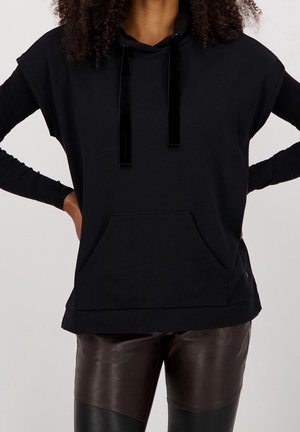 Personne portant un sweat à capuche noir à manches courtes avec poche avant et manches longues noires en dessous, associé à un pantalon en cuir marron foncé et noir.