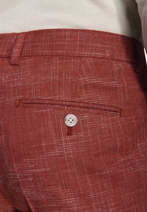 Nahaufnahme einer roten, strukturierten Stoffhose mit einer geknöpften Gesäßtasche und Gürtelschlaufen, getragen zu einem beigen Oberteil.