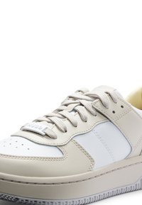 Beige sneakers med vita accenter. Slät läder- och textilmaterial, framträdande snörning, lätt perforering på tån, varumärkesetikett på sidan.