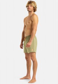 Zelene kopalne kratke hlače z elastičnim pasom in oranžno vrvico za zatezanje. Imajo majhno logotipno značko, enostavno teksturo in dolžino nad koleni.