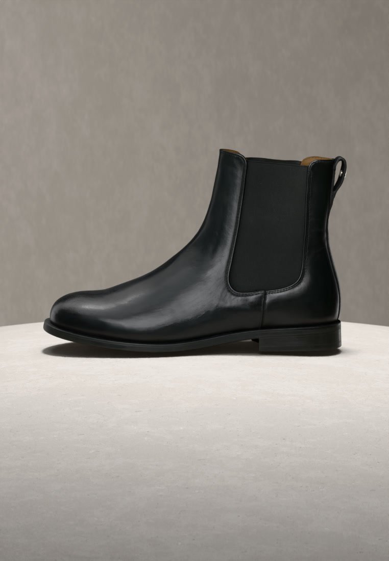 Bottine noire en cuir avec panneau latéral élastique et petit talon, posée sur une surface claire sur fond gris uni.
