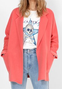 Korálový oversize blazer s klopami, bočními kapsami a uvolněným střihem, vrstvený přes grafické bílé tričko a džíny s vysokým pasem.