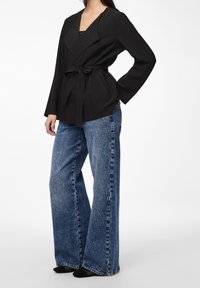 Veste ceinturée noire avec un col en V profond et des manches longues, associée à un jean denim bleu délavé à jambes larges. Tissu texturé, lignes épurées.