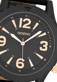 Montre noire avec un cadran rond, de grands chiffres beiges, des aiguilles en métal et un bracelet en caoutchouc. Présente une lunette texturée et un design minimaliste.