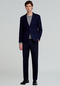 Blazer azul marino con un solo botón, combinado con un suéter gris y pantalones a juego. Zapatos de cuero negros completan el conjunto.