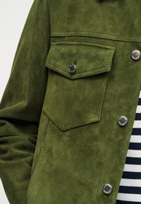 Κοντινό πλάνο ενός πράσινου σουέτ μπουφάν με τσέπη με καπάκι και μεταλλικά κουμπιά, φορεμένο πάνω από πουκάμισο με ναυτικό μπλε και λευκές ρίγες.