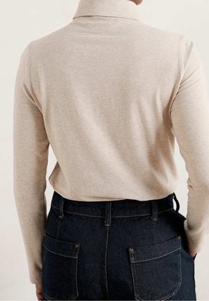 Haut beige à manches longues avec un col montant, associé à un jean en denim foncé avec des coutures dorées et des poches arrière. Texture de tissu lisse.